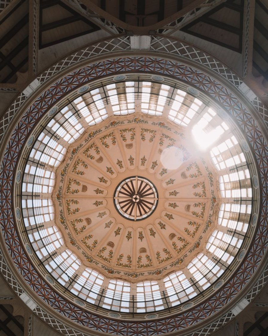 Interior del Mercado Central de Valencia bajo la cúpula de hierro y vidrieras, con puestos de fruta, embutidos y pescado y compradores entre los pasillos